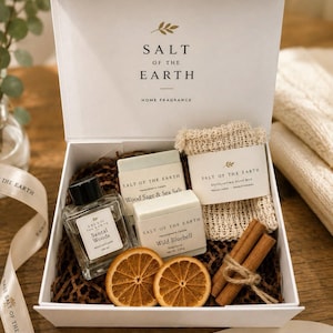May include: A white gift box labeled Salt of the Earth containing a glass bottle of Santal Woods reed diffuser, two soap bars labeled Wood Sage & Sea Salt and Wild Bluebell, an exfoliating soap bag, two dried orange slices, and two cinnamon sticks.