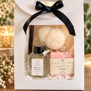May include: A white gift bag with a black ribbon bow containing a 100 ml English Pear & Freesia reed diffuser, a 100 g Rouge soap bar, and fluffy cream-coloured pom-poms. The labels read Salt of the Earth, Handcrafted in Canada.