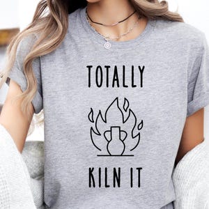 Puede incluir: Una camiseta gris jaspeado de manga corta con un estampado gráfico negro de un jarrón de cerámica rodeado de llamas. El texto "TOTALLY" está impreso sobre el gráfico y el texto "KILN IT" está impreso debajo del gráfico en una fuente simple.
