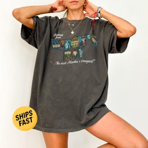 May include: A charcoal gray short-sleeve t-shirt featuring a graphic print that reads Greetings from WIDOW'S BAY ...the next Martha's Vineyard! The design includes multiple small rectangular images of people and a building in a collage style.