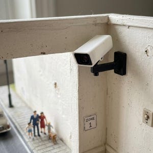 Puede incluir: Una cámara de seguridad en miniatura de color blanco montada sobre un soporte negro fijado a una pared blanca. Debajo de la cámara, un pequeño letrero cuadrado muestra el texto ZONE 1. La escena incluye una calle en miniatura con un coche.