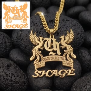 Puede incluir: Un collar con colgante dorado que presenta un monograma central flanqueado por dos aves fénix. Una cinta debajo dice SHAGE COLLECTIVE, Est. 2020, NYC, con la palabra SHAGE en letras grandes en la parte inferior. El colgante cuelga de una cadena dorada.