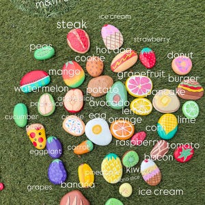 以下が含まれることがあります： m&m and blueberries, steak, ice cream, hot dog, strawberry, donut, watermelon, mango, cookie, potato, avocado, grapefruit, burger, pancake, lemon, lime, corn, cucumber, apple slice, egg, orange, pizza, carrot, raspberry, tomato, eggplant, bacon, grapes, banana, pineapple, kiwi, banana slice, breadなどの様々な食べ物が描かれた、滑らかな手描きの石のコレクション。