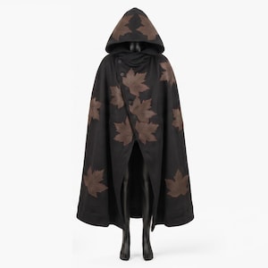 May include: A black hooded cloak featuring a pattern of brown maple leaf appliques scattered across the front and hood. The garment has a wide, flowing silhouette that drapes from the shoulders, with a pointed hood and a central opening.
