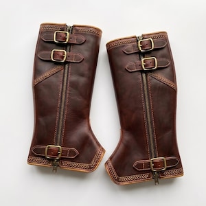 May include: A pair of dark brown leather gaiters featuring a central vertical zipper, three adjustable straps with brass-toned buckles, and decorative stitched chevron patterns along the top and bottom edges of each piece.