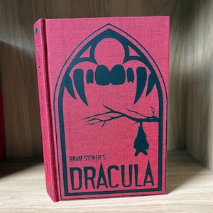 Może przedstawiać: Książka w twardej oprawie zatytułowana Bram Stoker's Dracula z czerwoną płócienną okładką. Z przodu znajduje się czarna grafika gotyckiego łuku z ostrymi kłami, nagą gałęzią drzewa i sylwetką wiszącego nietoperza. Tytuł jest wydrukowany na czarno.