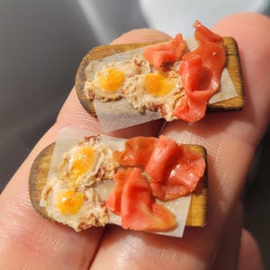 May include: Two miniature food-themed accessories featuring small wooden boards topped with translucent paper, realistic fried eggs with bright yellow yolks, and folded strips of pinkish-red cured meat, crafted with detailed textures.