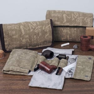 May include: A tan canvas tobacco pipe roll-up pouch featuring a dark brown script print and black trim. The open pouch reveals internal pockets containing two wooden tobacco pipes, metal pipe tools, and a small white cleaning brush accessory.