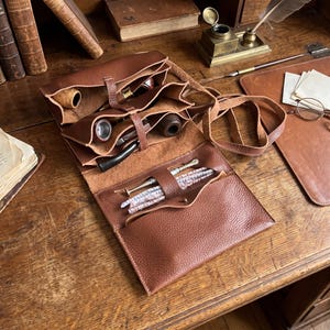 May include: A brown leather tobacco pipe roll with multiple interior compartments and a long strap. The roll contains several wooden smoking pipes, a metal tamper tool, and a collection of white and colored pipe cleaners stored in a front pocket.