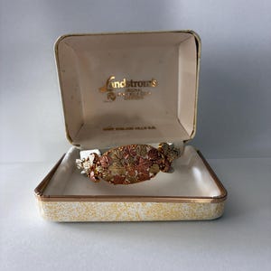 May include: A tri-colour gold bracelet featuring an oval centrepiece with detailed leaf motifs in yellow, rose, and green gold tones. The bracelet is presented in an open, cream-coloured Landstrom's jewellery box with gold lettering on the interior lid.
