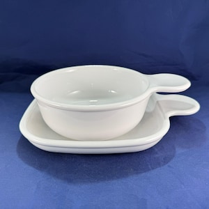 May include: A white ceramic soup bowl with a handle, resting on a matching saucer with a handle. The bowl and saucer are both round with a smooth, glossy finish. The set is on a blue background.