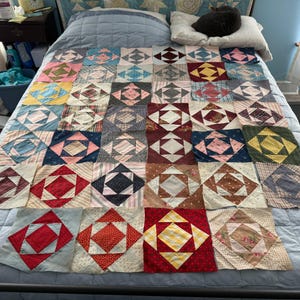 May include: A patchwork quilt in progress, featuring a diamond pattern in various colors and fabrics. The quilt blocks are arranged on a bed, showcasing a mix of floral, striped, and solid fabrics. The quilt is a handmade item.
