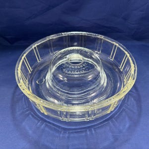 May include: Clear glass Queen Anne Glassbake bowl with a central raised section and a wide, ribbed outer rim. The raised section has a hole in the center. The text "Queen Anne Glassbake" is visible.