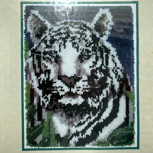 May include: A detailed needlepoint kit featuring a tiger's face. The artwork showcases a white tiger with black stripes, set against a dark blue and green background. The image is framed with a green border.