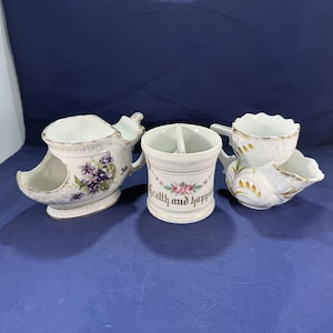 May include: Three ceramic shaving mugs with white surfaces and gold trim. The left mug features purple violet flowers. The center mug has a pink rose design and the text Health and happiness. The right mug displays a painted wheat pattern.