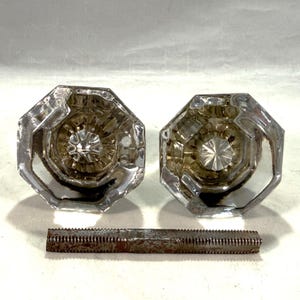 May include: Two clear glass door knobs with an octagonal shape and a faceted center design. The knobs feature a reflective surface and are positioned above a single, long, rectangular metal spindle with a threaded texture and visible rust.