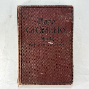 May include: A hardcover textbook titled Plane Geometry by Shutts, Revised Edition. The book features a textured, dark red fabric cover with black lettering and a thin black border frame on the front cover, showing signs of wear.
