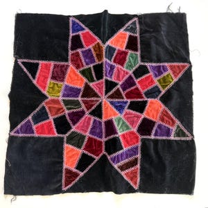 May include: A square fabric piece featuring a central eight-pointed star design composed of multicolored velvet patches in shades of red, purple, orange, and green, all joined together with visible white decorative zigzag stitching on black.