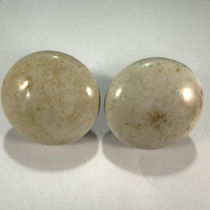 May include: Two round, off-white knobs with a speckled, aged appearance. The knobs have a glossy finish and are likely made of ceramic or a similar material. They are set against a plain white background.