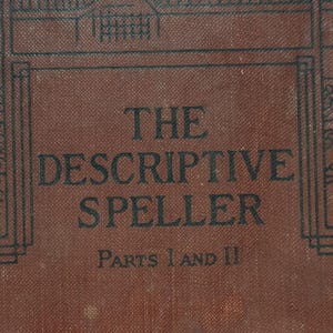 May include: A brown, textured book cover featuring the black printed text THE DESCRIPTIVE SPELLER PARTS I AND II. The cover includes a simple black line border design framing the title, with a visible woven fabric texture across the surface.