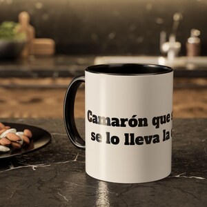 May include: A white ceramic mug with a black interior and a black handle. The exterior features bold black text that reads, "Camarón que se duerme se lo lleva la corriente," printed in a clear, sans-serif font on the side of the cylindrical vessel.