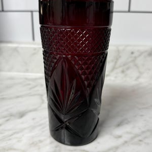 May include: A deep red glass tumbler featuring a textured pattern with a diamond-shaped grid at the top and a series of embossed leaf-like motifs around the lower section. The vessel has a cylindrical shape with a smooth rim and a solid base.