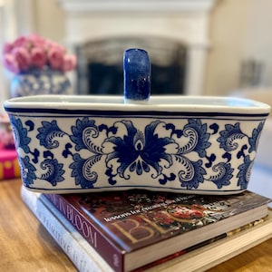 May include: A rectangular ceramic basket with a white base and a blue floral pattern. The container features a single arched handle in the centre, also decorated in blue. The exterior walls are adorned with symmetrical, intricate blue leaf motifs.