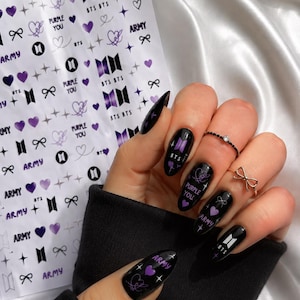 Peut inclure: Une planche de décalcomanies pour ongles avec des motifs BTS violets et noirs, incluant le logo BTS, le texte "PURPLE YOU" et "ARMY", des cœurs, des nœuds et des étoiles, avec des ongles peints en noir ornés de ces mêmes motifs.