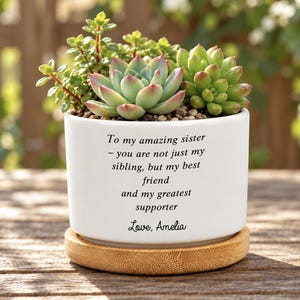 Puede incluir: Una maceta de cerámica blanca con un plato de bambú que contiene plantas suculentas verdes. La maceta tiene el texto impreso en negro: To my amazing sister - you are not just my sibling, but my best friend and my greatest supporter. Love, Amelia.