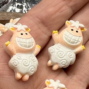 May include: Three smiling sheep-shaped embellishments. Each sheep has a light peach face and yellow accents on the ears and hooves. The sheep's body is white with a swirling pattern. The sheep are in a hand.