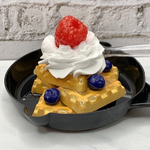 May include: A miniature waffle stack with whipped cream and a raspberry on top, topped with blueberries and a drizzle of syrup, served in a black skillet.
