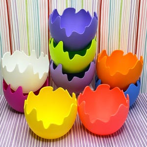 May include: A collection of colorful, decorative bowls in various shades, including purple, green, orange, and yellow. Each bowl has a unique, wavy rim design. The bowls are arranged on a striped background, showcasing their vibrant colors and shapes.