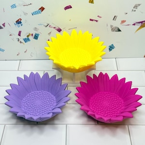 May include: Three colorful sunflower-shaped bowls in yellow, purple, and pink. The bowls have a textured center and petal design. The bowls are made of a solid material and are sitting on a white tiled surface.