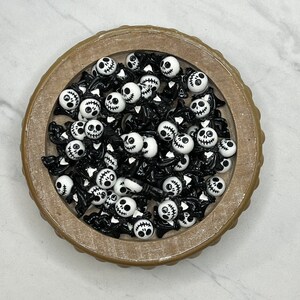 May include: A bowl filled with black and white skull-shaped charms. The charms have a stitched mouth and are perfect for adding a spooky touch to crafts.