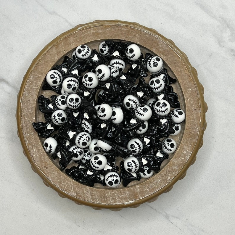 May include: A bowl filled with black and white skull-shaped charms. The charms have a stitched mouth and are perfect for adding a spooky touch to crafts.