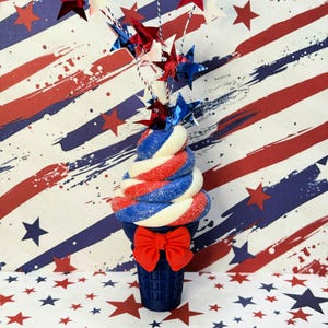 May include: A patriotic-themed decorative ice cream cone with red, white, and blue swirled layers, topped with star-shaped decorations. The cone is navy blue with a red bow, set against a star and stripe background.