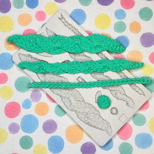 May include: A white mold with a braided design, featuring three bright green braided pieces. The mold also has a small green ball and two small circles. The background is a colorful polka dot pattern.