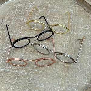 May include: A collection of wire-framed eyeglasses in gold, black, silver, and rose gold. The glasses have round lenses and thin temples. The eyeglasses are displayed on a light brown wooden surface.
