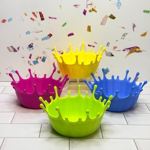 May include: A collection of five colorful, splash-shaped bowls in bright hues of pink, yellow, blue, and green. The bowls are arranged on a white tiled surface, with confetti in the background.
