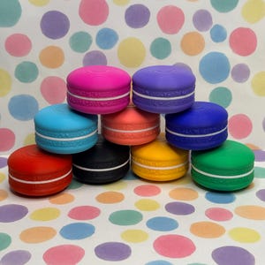 May include: A collection of colorful, round objects resembling macarons. The objects are stacked in a pyramid shape, with colors including pink, purple, blue, orange, black, yellow, and green. Each has a white band around the middle.