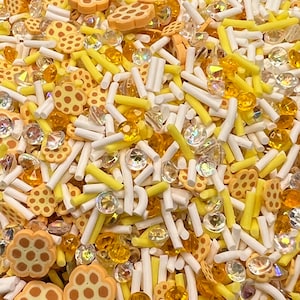 May include: A close-up of a mixture of yellow and white sprinkles. The sprinkles are in the shape of honeycomb, diamonds, and long, thin sticks.