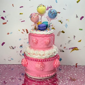May include: A three-tiered pink cake with white frosting and decorative pink roses. The cake is topped with colorful cake pops and macarons. Confetti is scattered around the cake, creating a festive atmosphere.