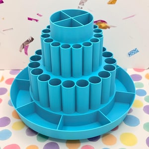 May include: A three-tiered, light blue organizer with multiple cylindrical compartments and trays. The top tier has a divided section, while the lower tiers feature open storage for art supplies or office items. The organizer is made of plastic.