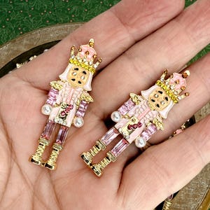 May include: Two pink and gold nutcracker earrings with crowns and pearl accents. The earrings have a detailed design with pink, gold, and clear crystal accents. The earrings are in the shape of a nutcracker soldier.