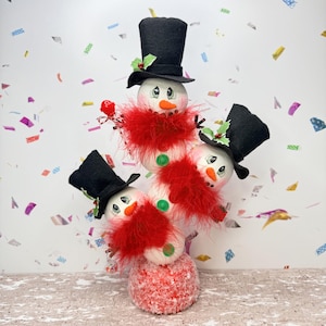 May include: A whimsical snowman decoration featuring four snowmen stacked on top of each other. Each snowman has a black top hat, a red feather boa, and a carrot nose. The base is a red and white textured ball.