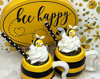 May include: Two yellow and black striped mugs with bee designs and white whipped cream on top. The mugs have black and white striped straws. A yellow oval sign with the text "bee happy" is in the background.