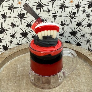 May include: A clear glass mug filled with layers of red and black liquid, topped with a red and white vampire teeth decoration and a small knife. The background features a spider web pattern.