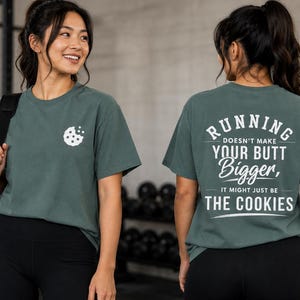 May include: A dark sage green short-sleeve t-shirt featuring a small white cookie graphic on the front left chest and white text on the back that reads: RUNNING DOESN'T MAKE YOUR BUTT Bigger, IT MIGHT JUST BE THE COOKIES, with decorative lines.
