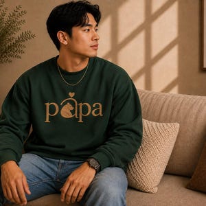 May include: A dark green long-sleeve sweatshirt featuring the word papa in gold lettering across the chest. The letter p incorporates a small bear graphic and a heart symbol above it. The sweatshirt has a crew neckline and ribbed cuffs.