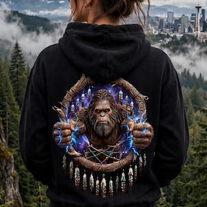 May include: A black hooded sweatshirt featuring a large graphic on the back. The design shows a brown Bigfoot creature emerging from a circular, wooden-textured frame decorated with hanging feathers and beads, surrounded by blue lightning.